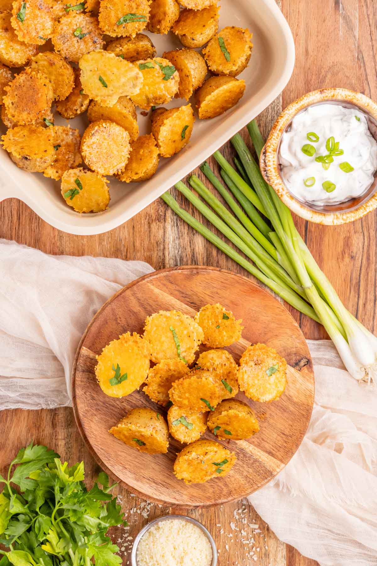 A wooden plate filled with crispy parmesan roasted potatoes with the pan of potatoes in a red serving dish in the background.