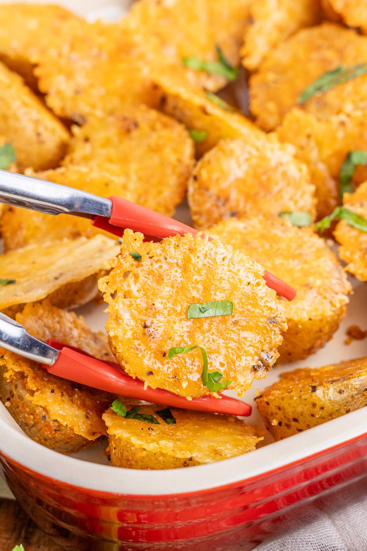 Tongs removing a parmesan roasted potato from a red dish.