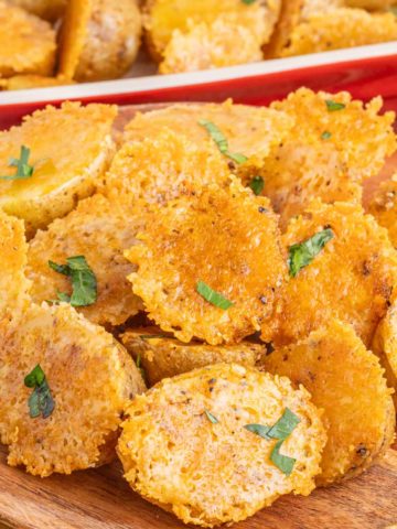 A wooden plate filled with crispy parmesan roasted potatoes with the pan of potatoes in a red serving dish in the background.