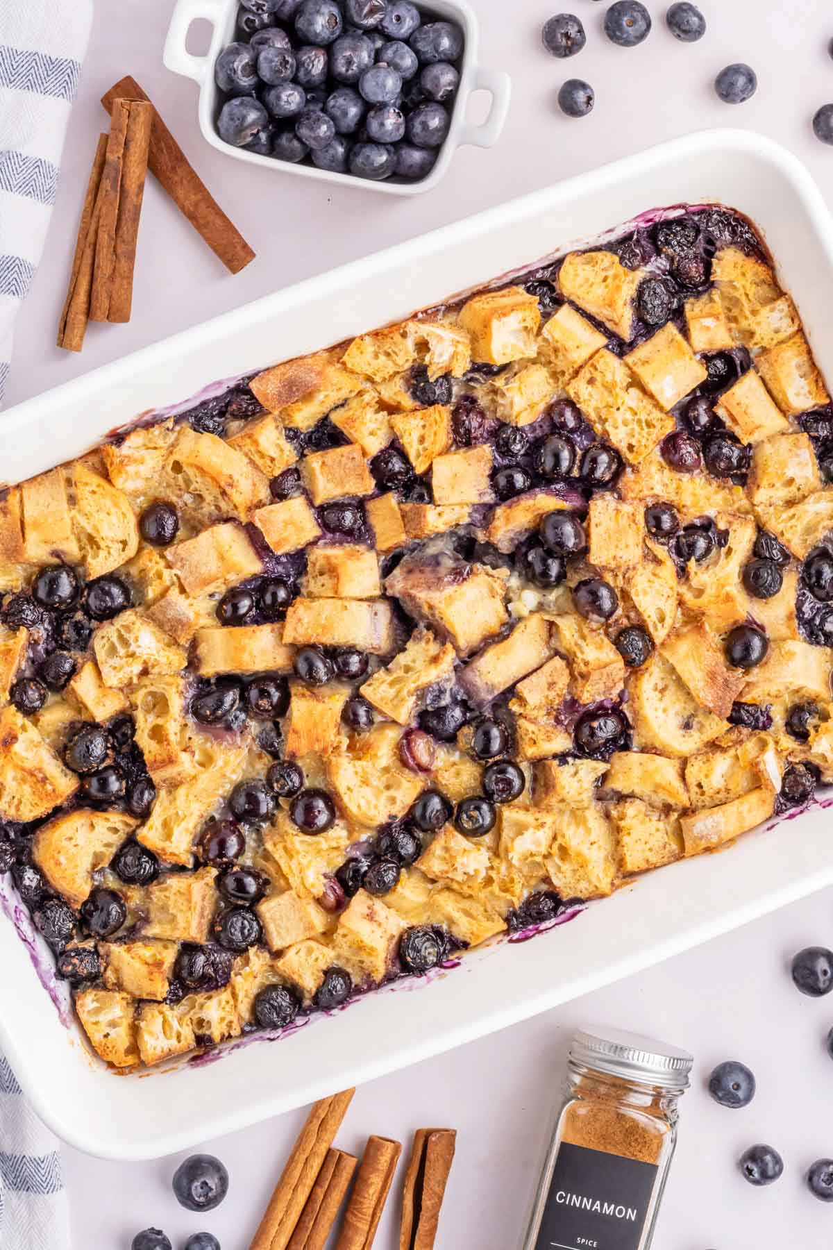 A white casserole dish filled with blueberry french toast casserole on a white table with a container of blueberries and cinnamon sicks set in the backdrop.