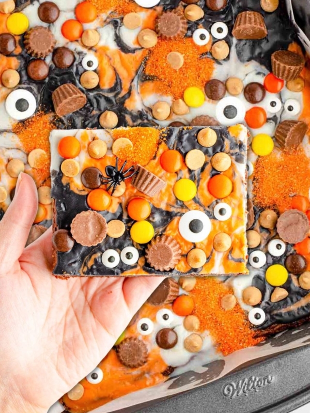 A hand holding a serving of the Halloween Brownies- and a tray in the background of the iced peanut butter brownies topped with candies, peanutbutter cups and candy eyeballs.