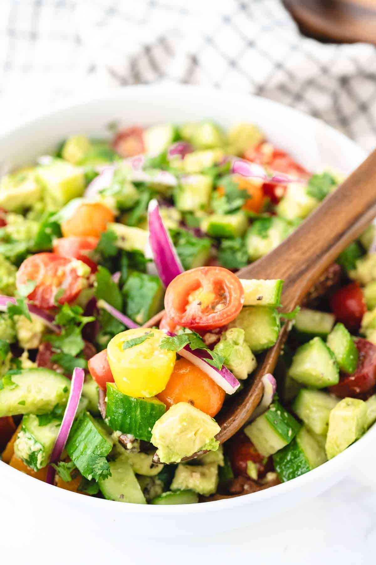 A wooden spoon removing a scoop of avocado salad with cucumbers, tomatoes, and onions.