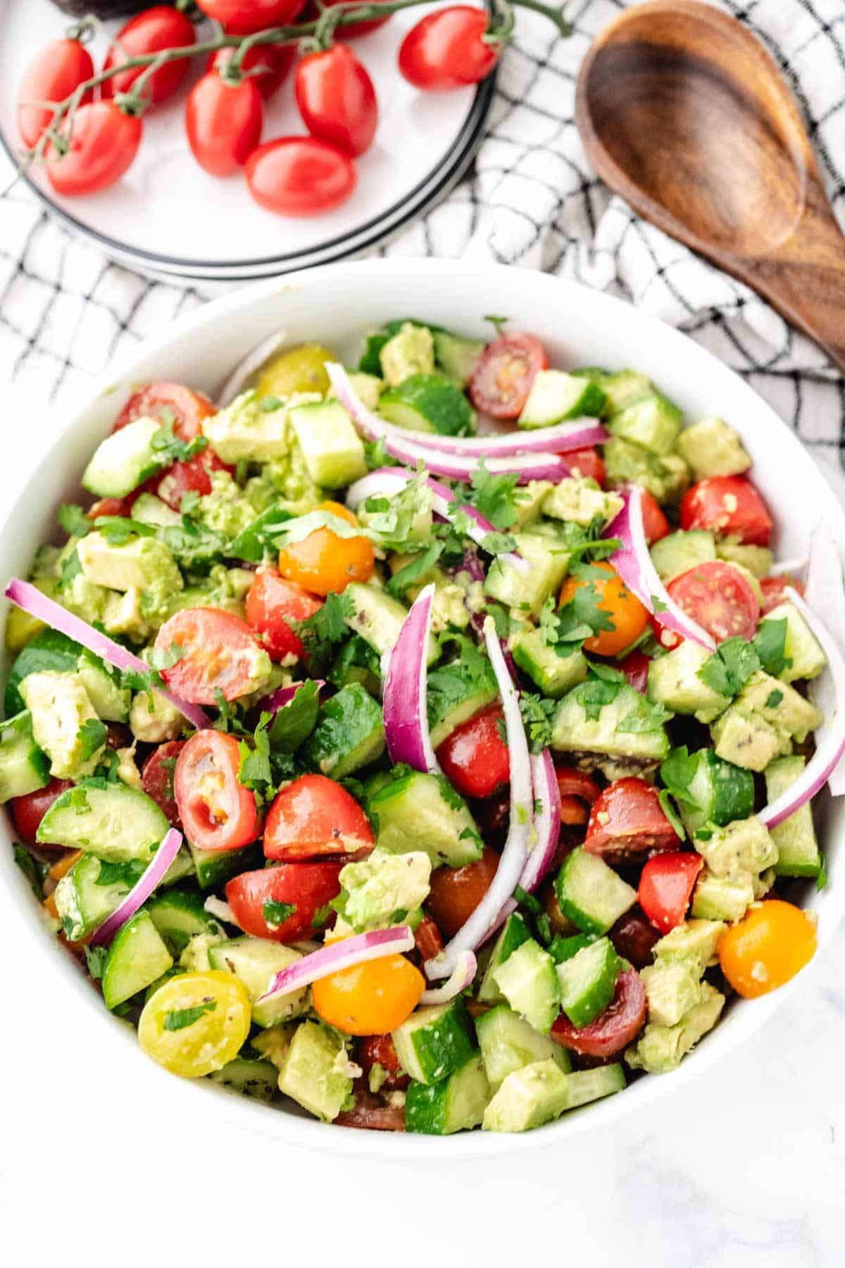 A cucumber tomato avocado salad tossed in a lemon vinaigrette dressing in a white serving bowl with a wooden spoon in the background.