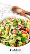 A close up centered image of the cucumber tomato salad with avocado and wooden serving spoons in the distance.