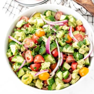 A cucumber tomato avocado salad tossed in a lemon vinaigrette dressing in a white serving bowl.