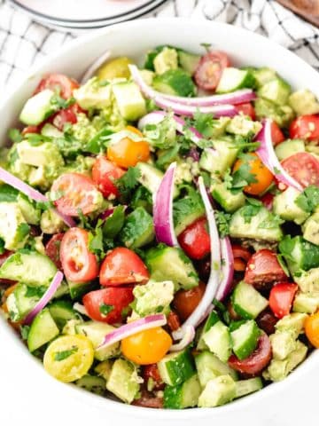 A cucumber tomato avocado salad tossed in a lemon vinaigrette dressing in a white serving bowl.