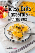 A white plate with black rim with a serving of cheesy grits casserole with breakfast sausage set on a table with fruit and the casserole dish in the background.