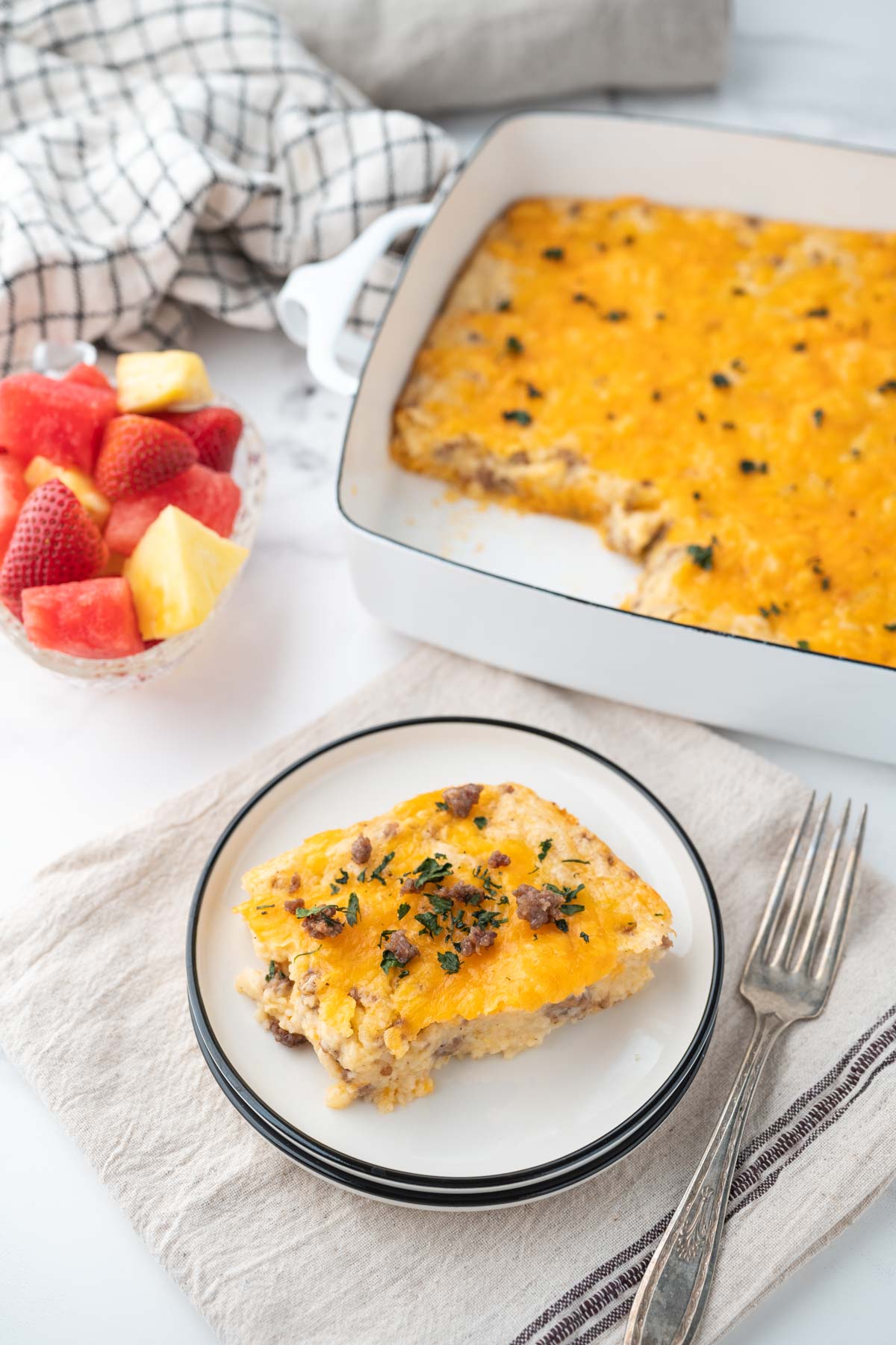 A slice of cheese grits and sausage casserole with the serving dish and a bowl of fruit on the table.