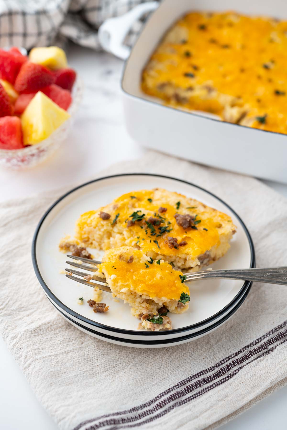 A white plate with black rim with a serving of cheesy grits casserole with breakfast sausage set on a table with fruit and the casserole dish in the background.