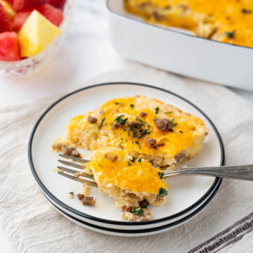 A white plate with black rim with a serving of cheesy grits casserole with breakfast sausage set on a table with fruit and the casserole dish in the background.