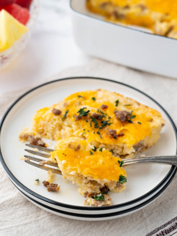 A white plate with black rim with a serving of cheesy grits casserole with breakfast sausage set on a table with fruit and the casserole dish in the background.