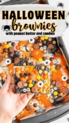 A hand holding a serving of the Halloween Brownies- and a tray in the background of the iced peanut butter brownies topped with candies, peanutbutter cups and candy eyeballs.
