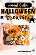 A hand is holding out a slice of the Peanut Butter Halloween Brownies with icing, candies, chips and candy eyeballs.