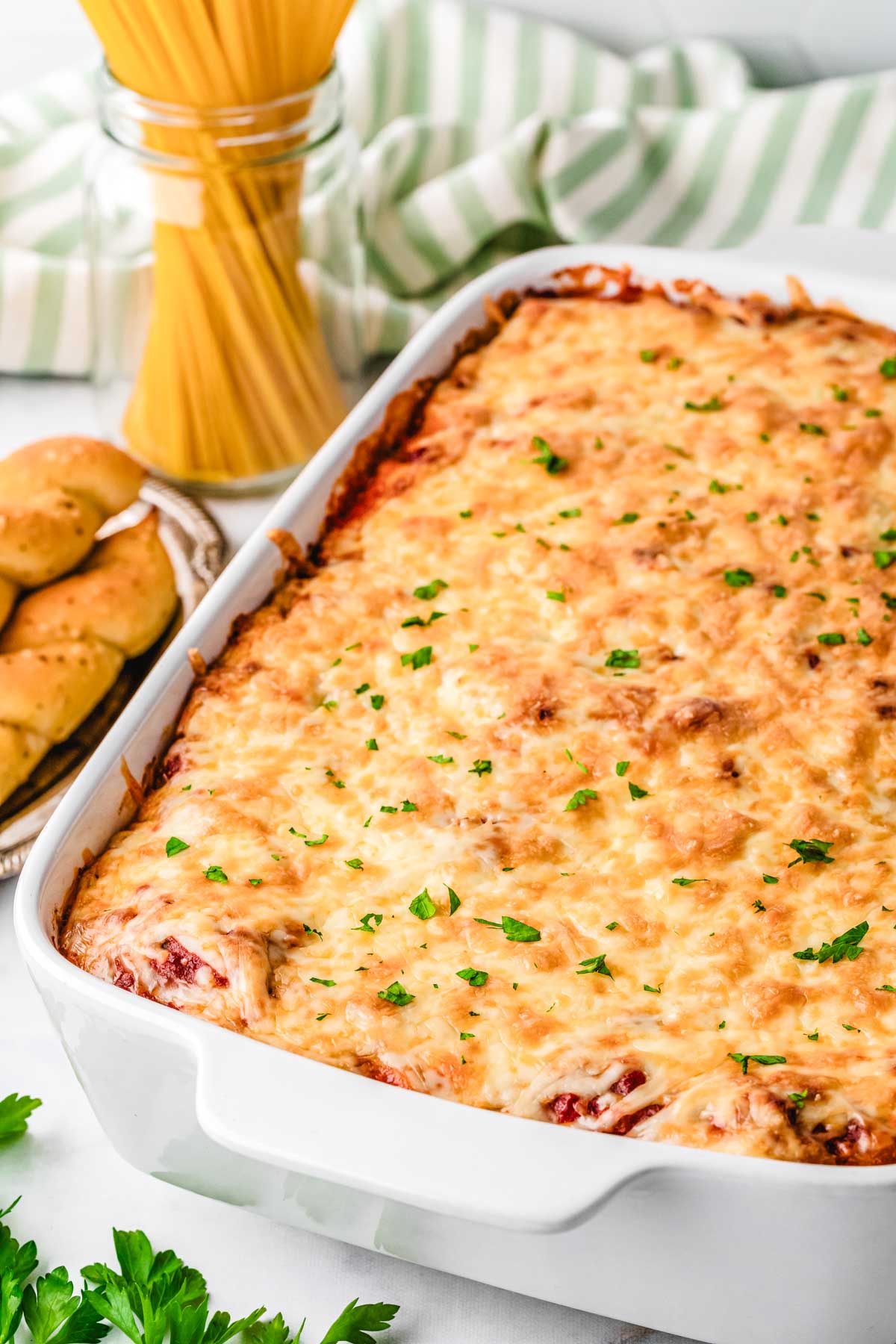 A pan of ultimate baked spaghetti set on a table with breadsticks and a green and white tea towel.