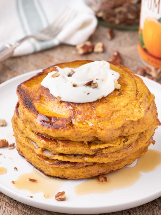 A stack of pumpkin pancakes on a white plate set on a wooden table.