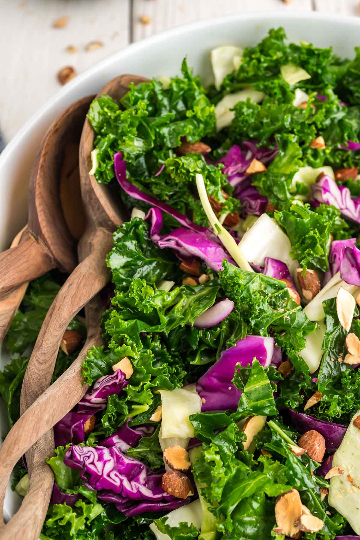 A close up of kale crunch salad in a white serving bowl with wooden serving spoons to the side.