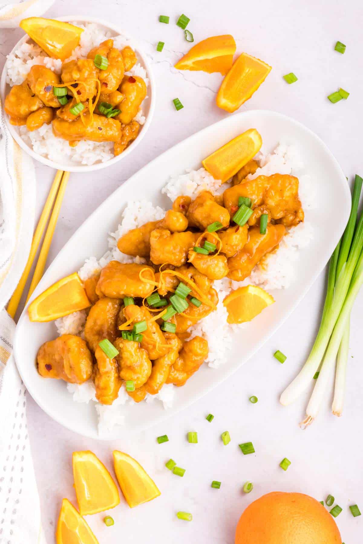 A white table set with a serving plate of Orange chicken on top of white rice decorated with orange slices and sliced green onions.