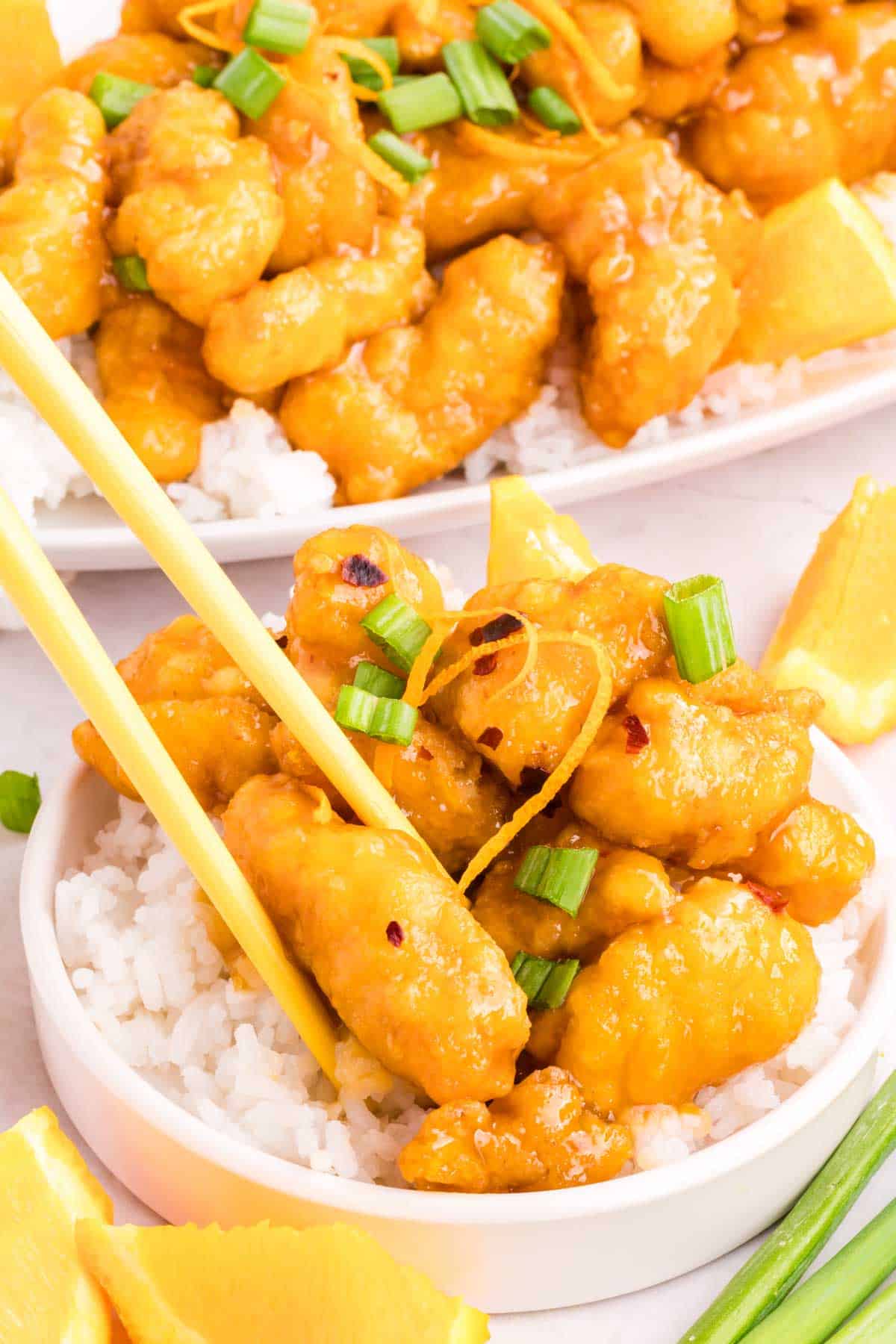 Chopsticks picking up a piece of orange chicken from a white plate with chinese orange chicken on top of white rice.