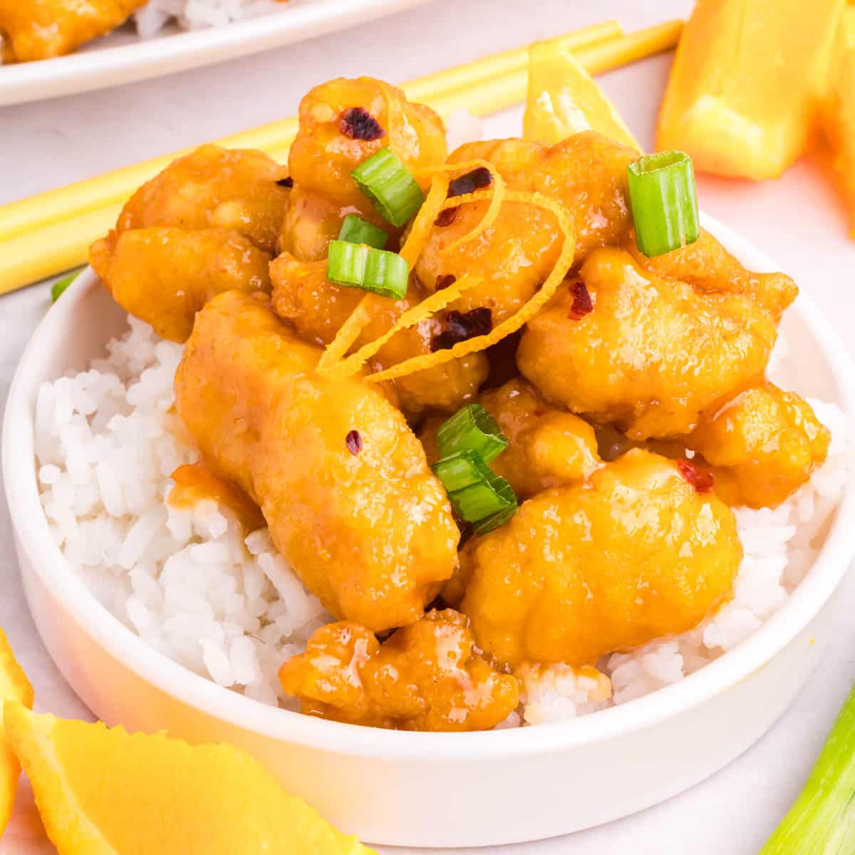 Chopsticks picking up a piece of orange chicken from a white plate with chinese orange chicken on top of white rice.