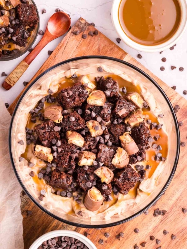 A layered brownie dessert with chocolate cheesecake filling and caramel in a serving dish on a wooden tray.