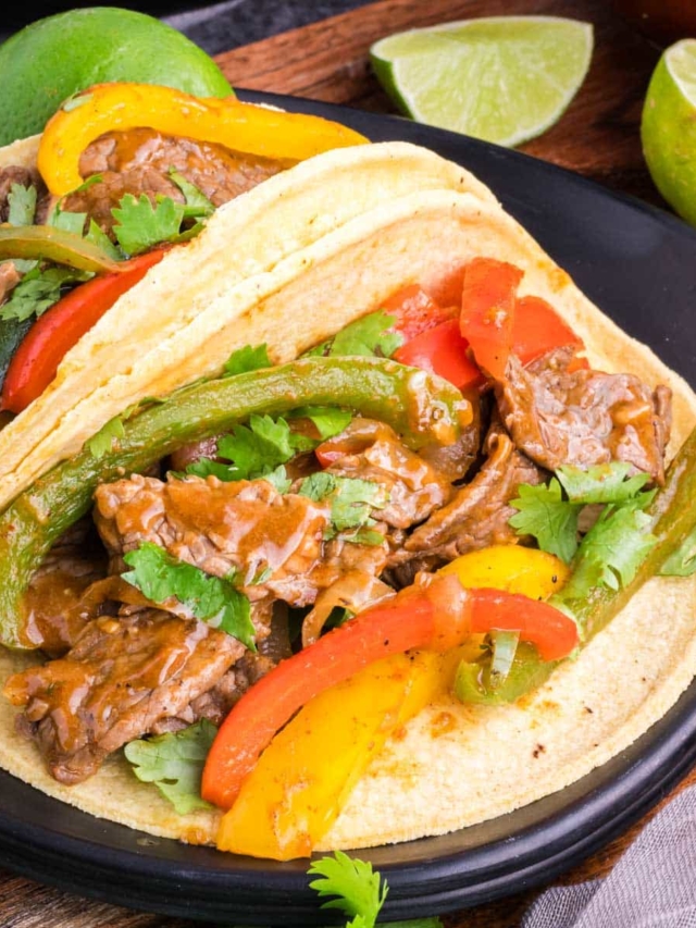 Two steak fajitas served in corn tortillas on a black plate.