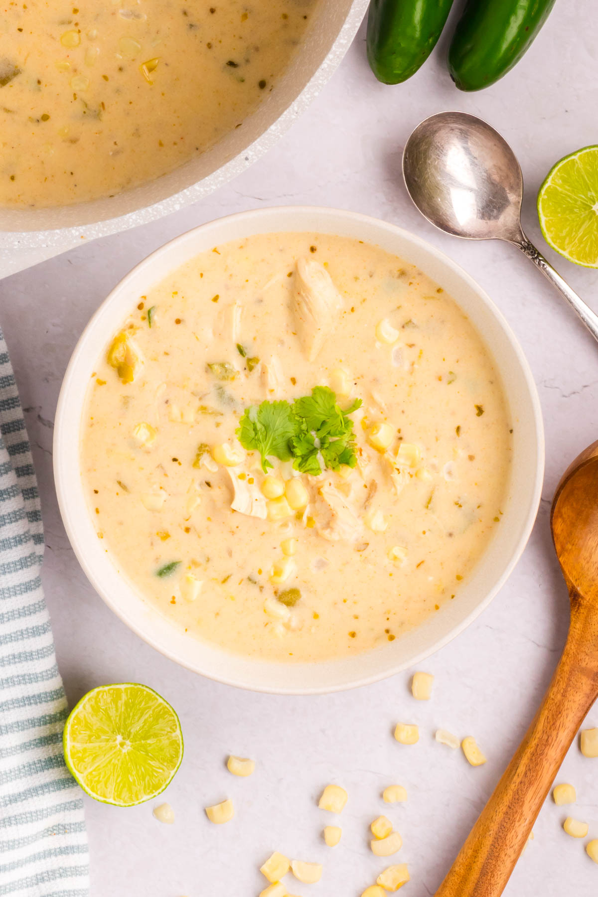 A bowl of Mexican street corn white chicken chili on a table with corn kernels, lime, and cilantro.