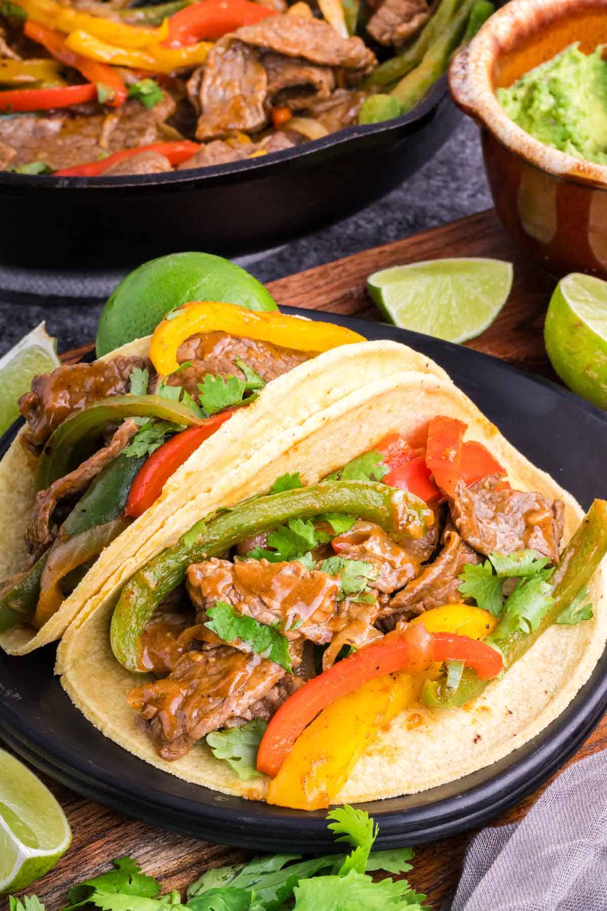 Two steak fajitas served in corn tortillas on a black plate.