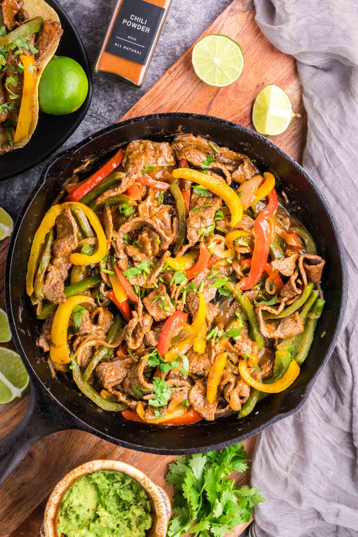 Steak fajitas with onions and peppers in a black cast iron skillet set on a table with lime, cilantro, and guacamole.