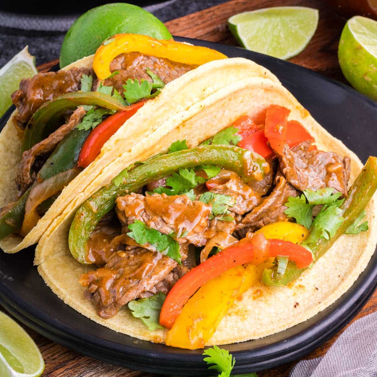 Two steak fajitas served in corn tortillas on a black plate.