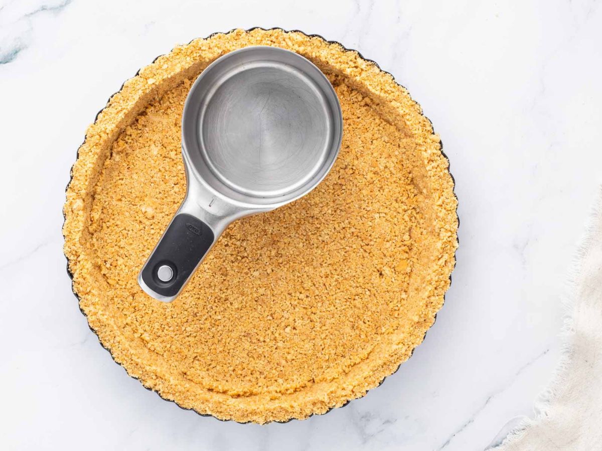 A pie crust being pressed into pan with a measuring cup.