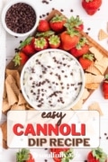 A cutting board with a bowl of easy cannoli dip topped with chocolate chips and surrounded by strawberries and bits of sugar cone.