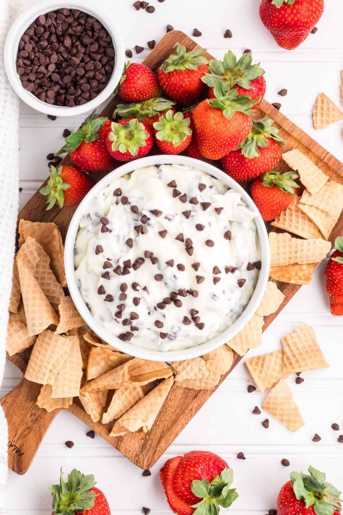 Easy Cannoli Dip in a white bowl served on a wooden serving board with broken waffle cones and strawberries.