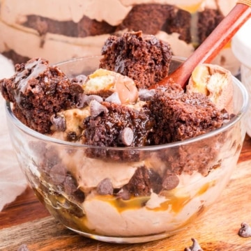 A clear dessert bowl will a layered chocolate cheesecake brownie trifle with caramel sauce and Twix candy bar topping.