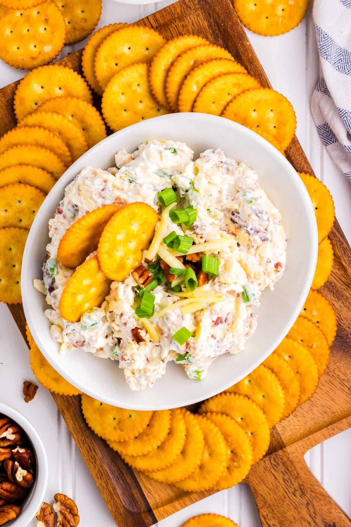 A wooden tray with a white bowl filled with million dollar dip surrounded by Ritz crakers.