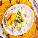 A wooden tray with a white bowl filled with million dollar dip surrounded by Ritz crakers.