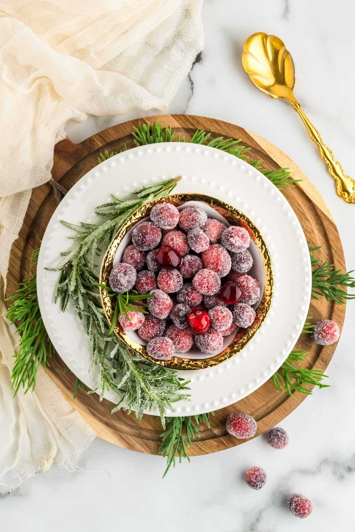 Sugared cranberries in a gold rimmed bowl set on a white charger and wooden board garnished with sugared rosemary.