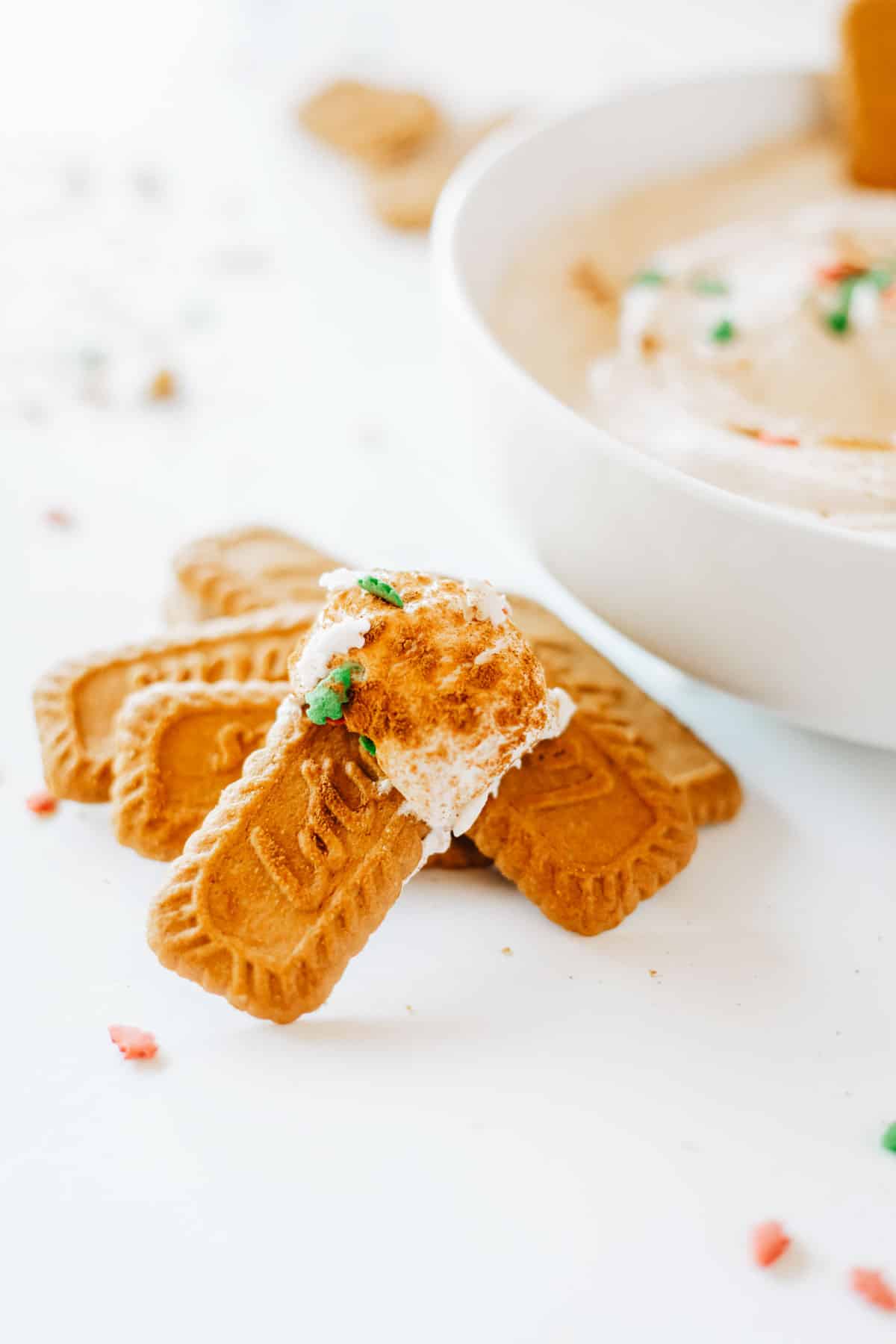 A biscoff cookie dipped into eggnog dip and set on a white table with a stack of cookies.