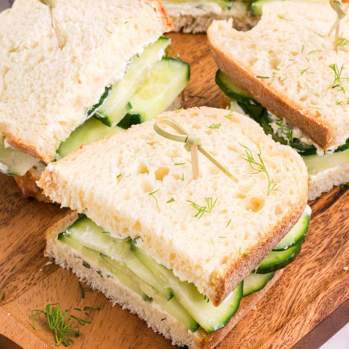 Sliced cucumber sandwiches on a wooden tray with dill sprinkled on top.