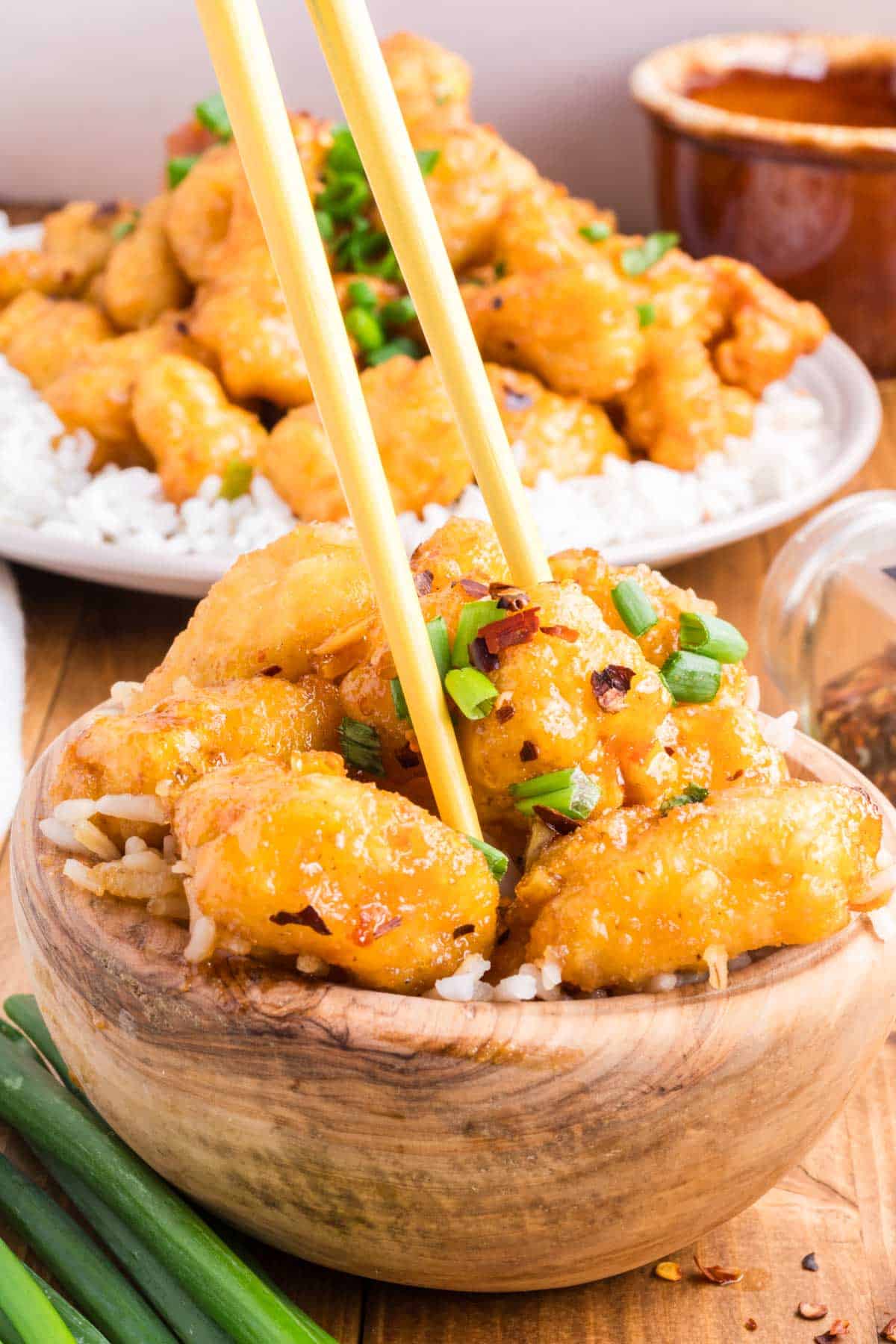A wooden bowl filled with honey garlic chicken with rice being picked up with chopsticks.