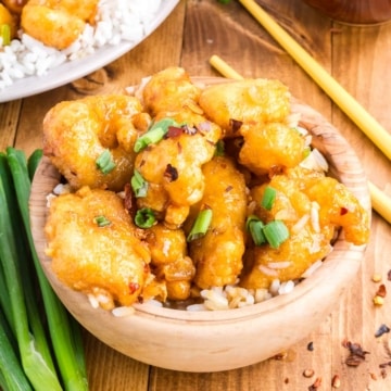 A light wooden bowl filled with rice and topped with crispy honey garlic chicken garnished with sliced green onions, set on a wooden table with a bunch on spring onions and red pepper flakes.
