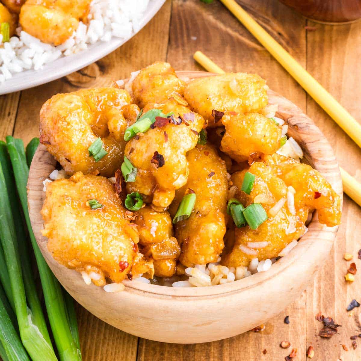 A light wooden bowl filled with rice and topped with crispy honey garlic chicken garnished with sliced green onions, set on a wooden table with a bunch on spring onions and red pepper flakes.