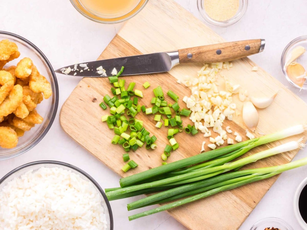 A wooden cutting board with a knife, green onions, and garlic cloves diced up.