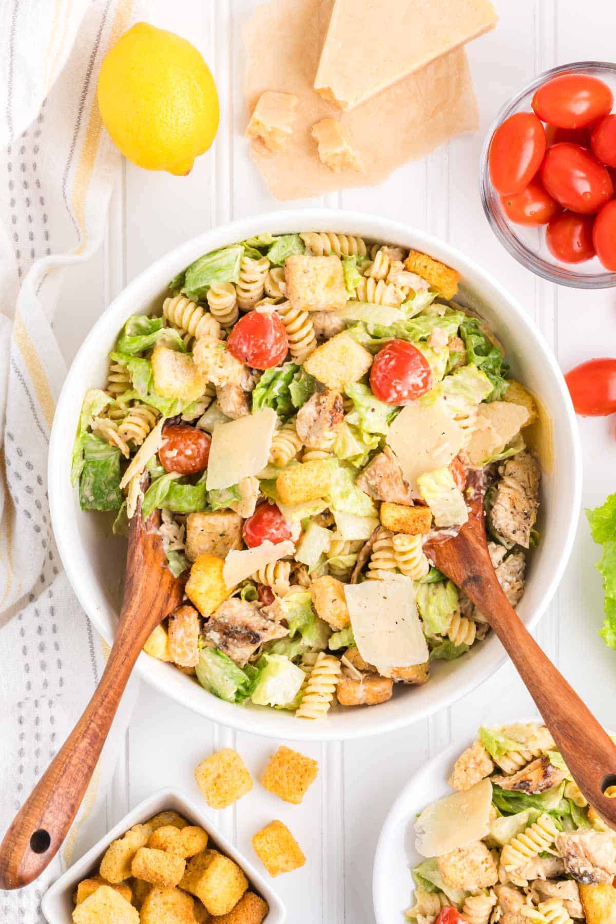 A chicken caesar pasta salad in a white bowl being served with wooden spoons.