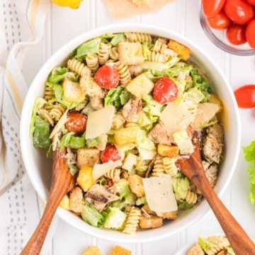 A chicken caesar pasta salad in a white bowl being served with wooden spoons.