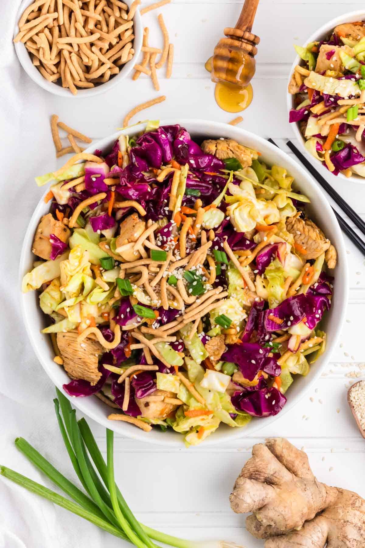 A Chinese Chicken Salad on a white wood tablescape surrounded by green onion, ginger, sesame seeds, and crunchy noodles.