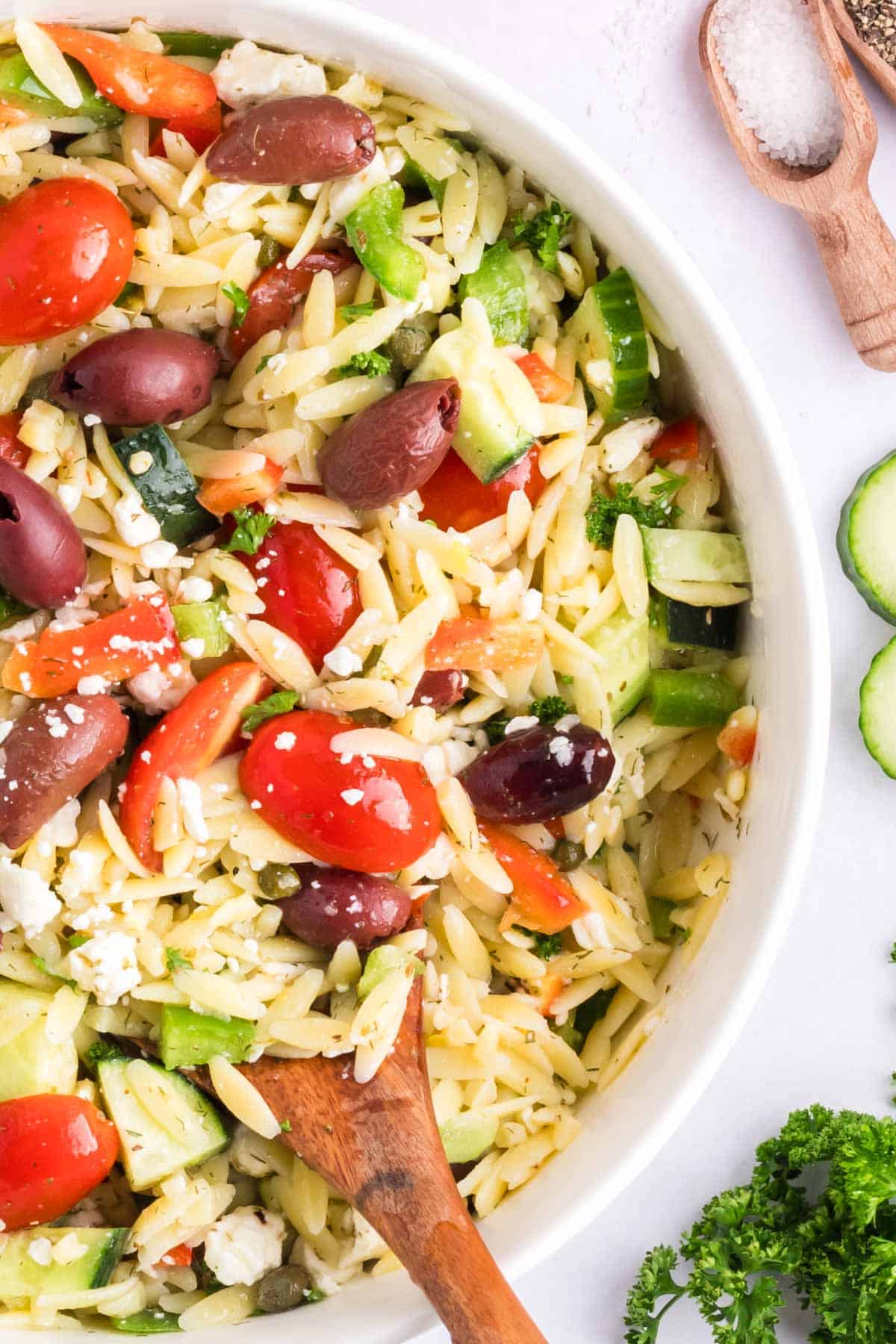 A white serving bowl with orzo salad and a wooden serving spoon.