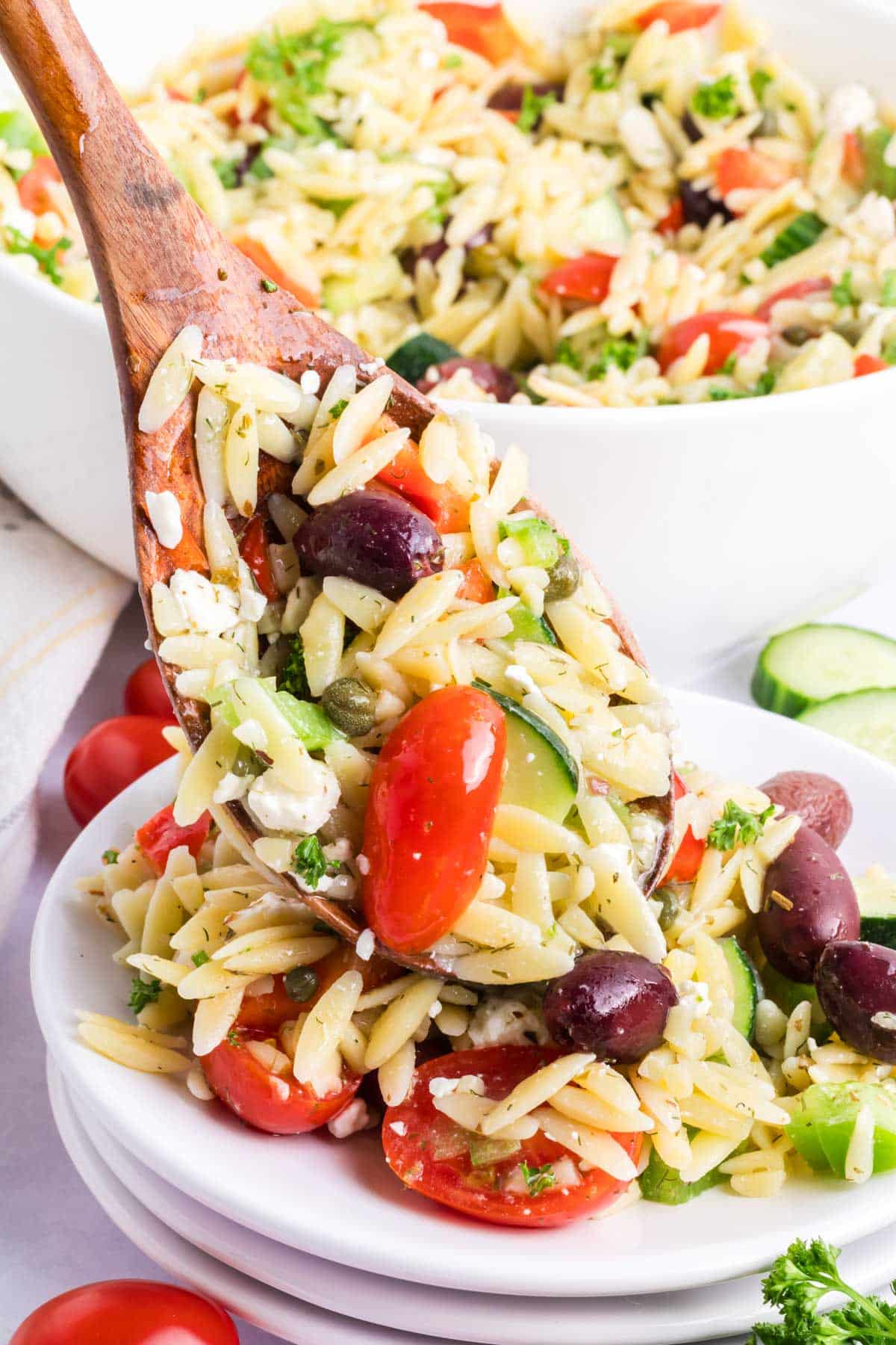 A wooden spoon putting Greek orzo salad on a white plate.