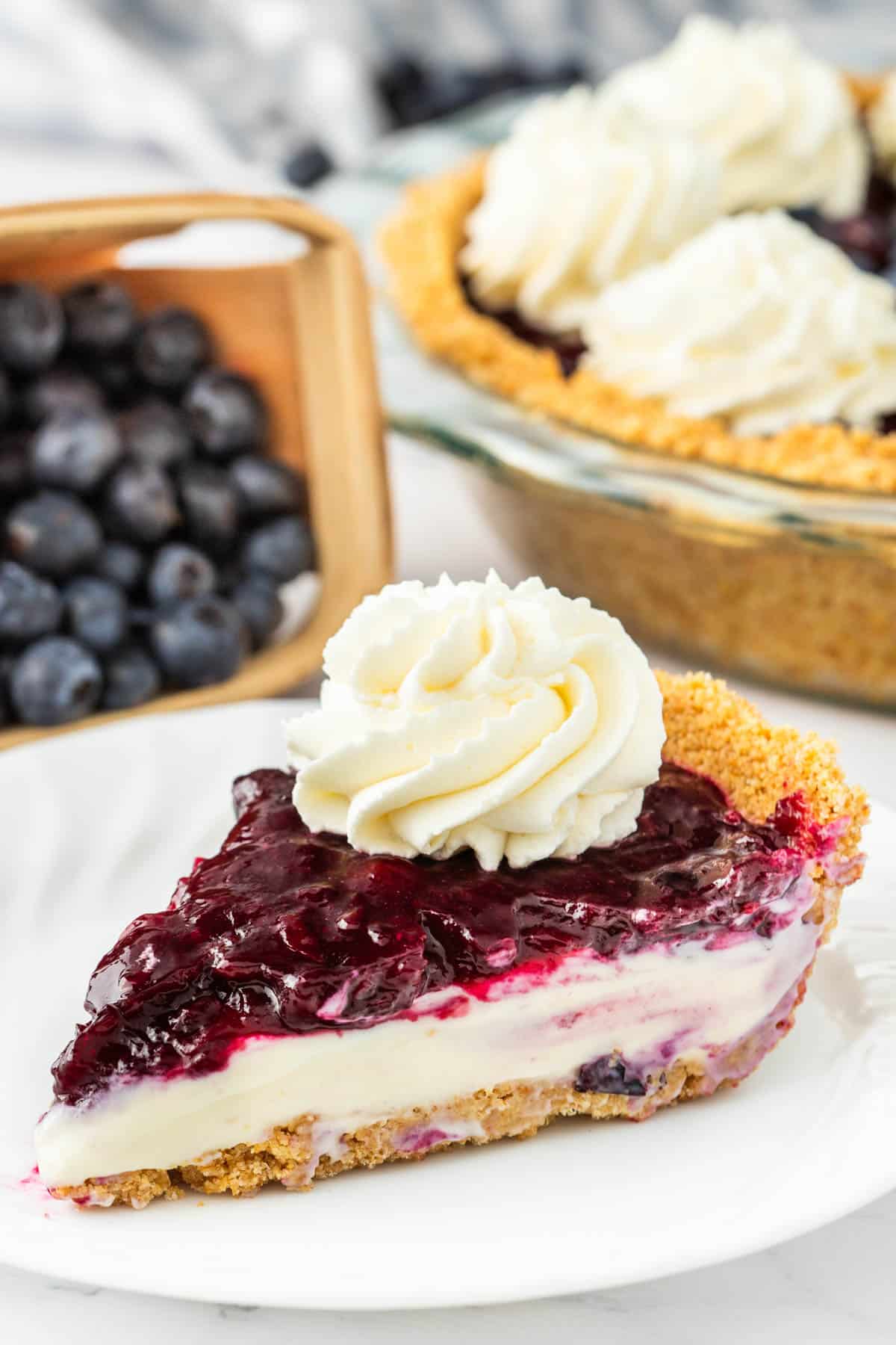 A slice of blueberry cheesecake pie on a white plate with the whole pie in the background.