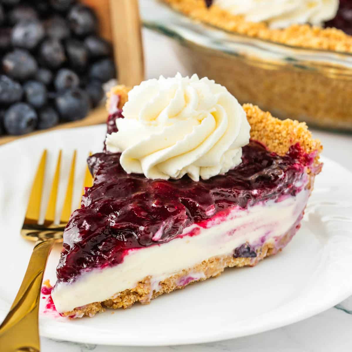 A slice of blueberry cheesecake pie on a white plate with a gold fork to the side of the pie.