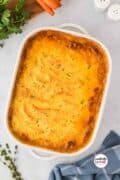 A whole shepherd's pie in a white casserole dish on a table with a sprig of thyme and a blue tea towel beside it.